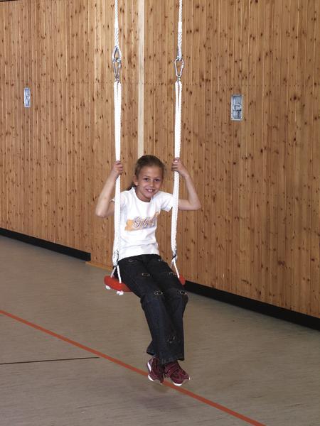 Kind auf Schaukel in Turnhalle mit Holzverkleidung, lächelnd und spielend.