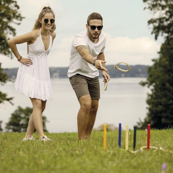 Junges Paar spielt Ringwurfspiel im Freien mit See und Bäumen im Hintergrund.