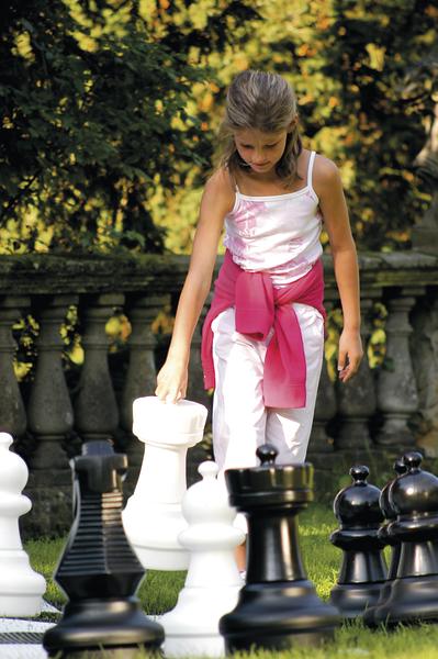 Mädchen spielt im Garten mit übergroßen Schachfiguren auf grünem Rasen.
