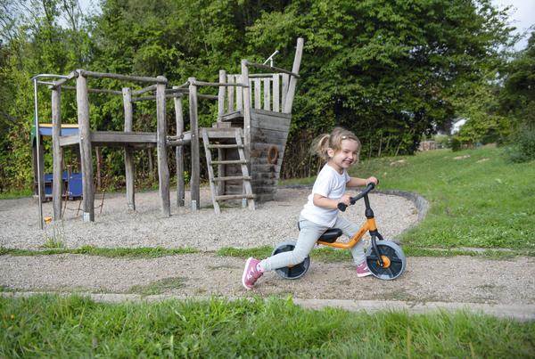 Kind auf Laufrad spielt auf Spielplatz mit Klettergerüst und Rutsche im Grünen.