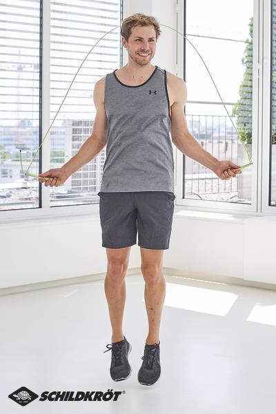 Mann in Sportkleidung springt mit Springseil in hellem Raum vor Fenster.