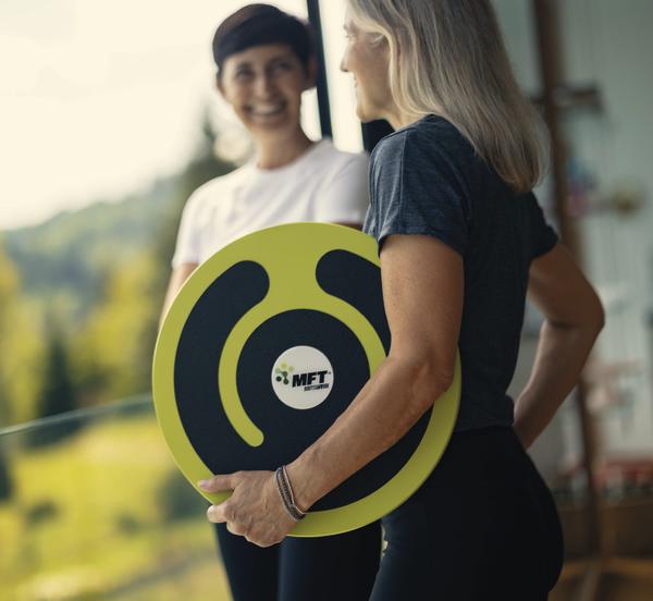 Frau hält MFT Balance-Board, spricht mit Trainerin im Fitnessstudio mit Naturblick.