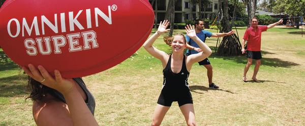 Gruppe spielt im Park mit großem roten OMNIKIN Super Ball bei sonnigem Wetter.
