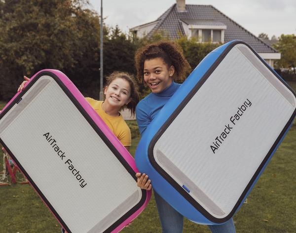 Zwei Mädchen halten bunte AirTrack Factory Luftturnmatten im Garten vor einem Haus.