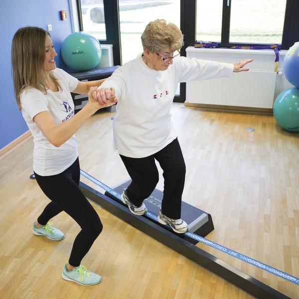 Ältere Frau trainiert Gleichgewicht auf Slackline mit Unterstützung einer Trainerin im Fitnessraum.