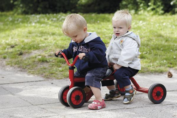 Zwei Kleinkinder fahren gemeinsam auf einem roten Dreirad im Park.