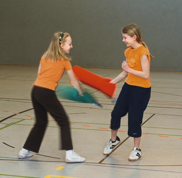 Zwei Mädchen in Sportkleidung kämpfen spielerisch mit Schaumstoffschlägern in der Turnhalle.