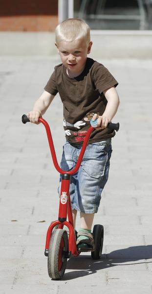 Kleiner Junge fährt mit rotem Dreirad auf gepflastertem Weg bei Sonnenschein.