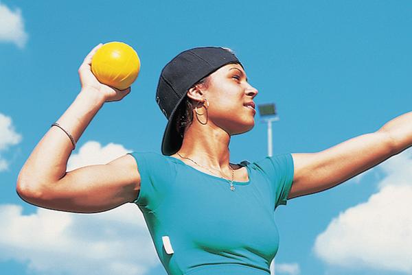 Frau mit schwarzer Kappe wirft gelben Ball bei sonnigem Himmel im Freien.