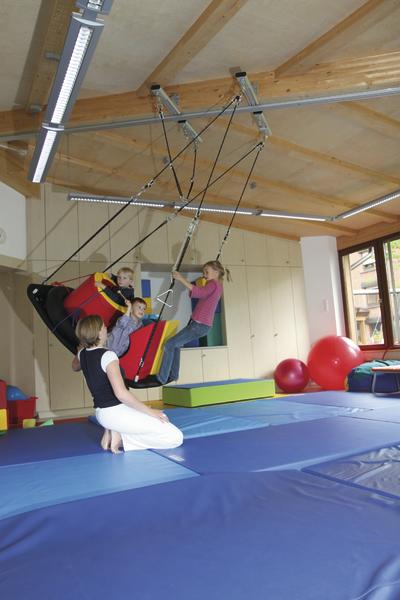 Kinder spielen auf einer Therapieschaukel in einem hellen Bewegungsraum mit Matten und Gymnastikbällen.
