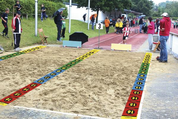 Kinderwettkampf im Weitsprung auf einer Sportanlage mit Zuschauern und Betreuern.