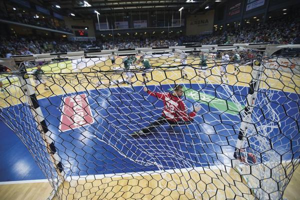 Torwart in rotem Trikot hält Ball im Handballspiel vor vollem Publikum in Halle.
