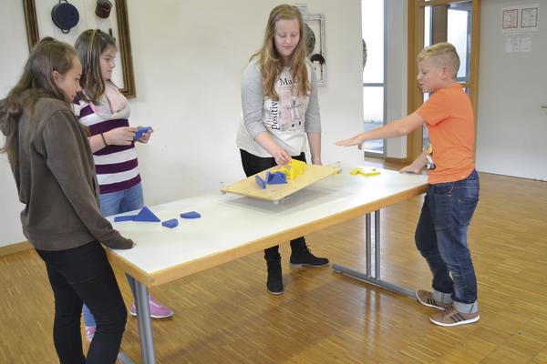 Kinder spielen ein Balancespiel mit Holzbrett und geometrischen Formen in einem Klassenraum.