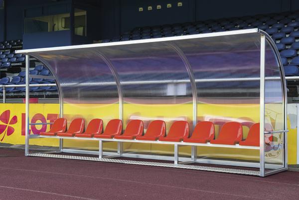 Überdachte Ersatzbank mit roten Sitzen im Fußballstadion vor leeren Tribünen.