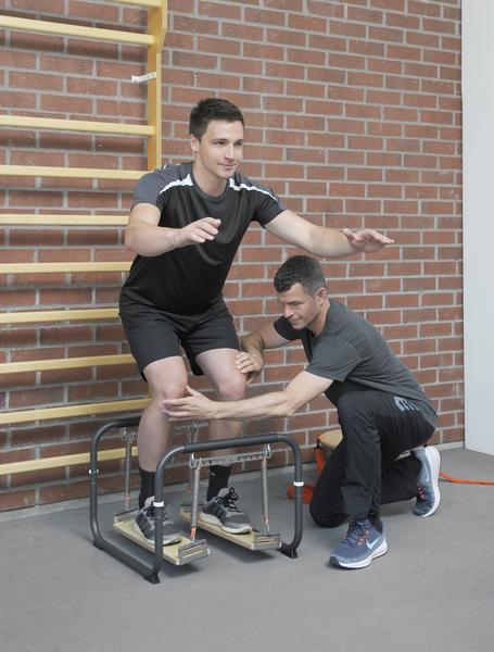 Junger Mann macht Balance-Übung auf Wackelbrett, Trainer unterstützt ihn in Sporthalle.