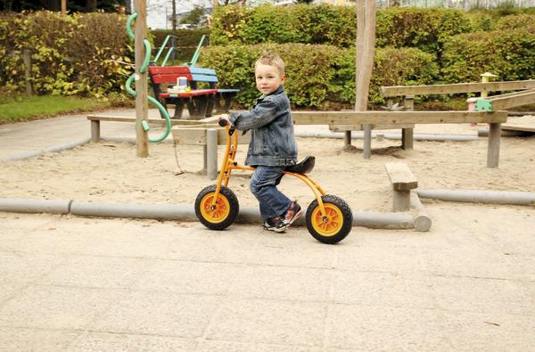 Kind auf orangem Laufrad im Spielplatz mit Sand und Klettergerüst im Hintergrund.