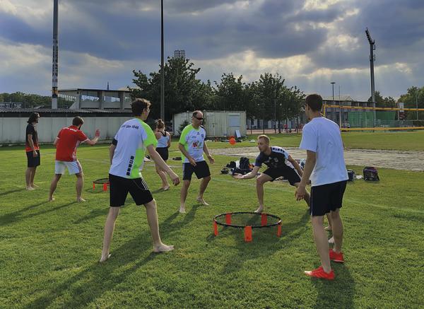 Gruppe junger Männer spielt Spikeball auf einer grünen Wiese bei bewölktem Himmel.