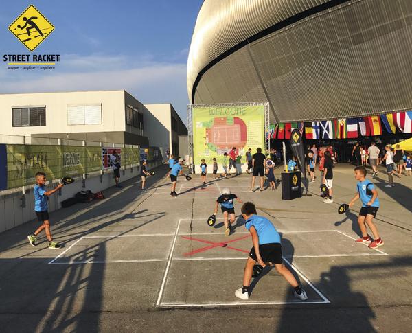 Kinder spielen Street Racket draußen vor einer großen Sportarena mit internationalen Flaggen.