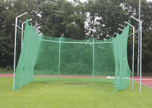 Hammerwurfanlage mit grünem Schutznetz auf Sportplatz vor Waldkulisse.