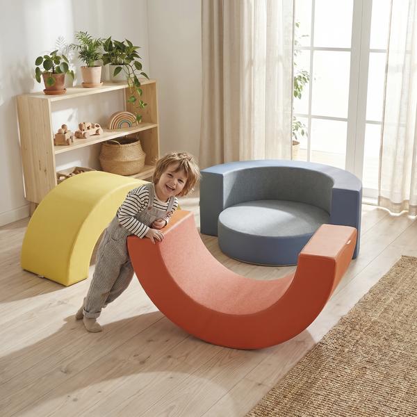 Anwendungsbild eines Kindes beim Spielen mit gebogenen Hüpfkissen-Modulen in einem hellen Spielzimmer. Im Hintergrund sind ein rundes Hüpfkissen mit Rückenlehne in Blau sowie eine gelbe, gebogene Rückenlehne zu sehen.