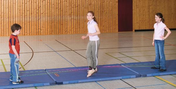 Kinder springen in einer Turnhalle auf einer Matte beim Seilspringen.