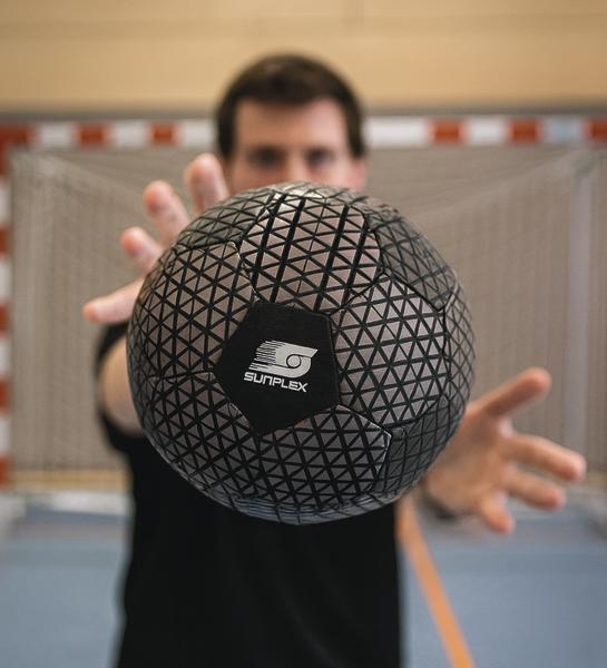 Mann hält einen Ball in dunklem Braun mit schwarzem Muster und Sunflex-Logo vor sich.