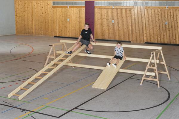 Kinder spielen auf einer Holzrutsche und Klettergerüst in einer Turnhalle.