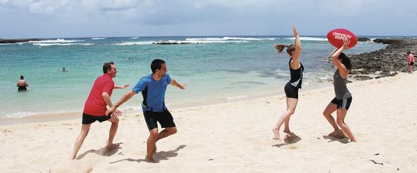 Vier Personen spielen Beachball am Sandstrand mit klarem Meer und bewölktem Himmel im Hintergrund.
