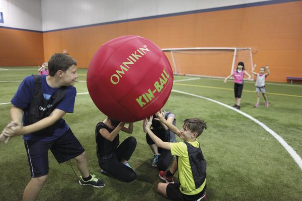 Kinder spielen Kin-Ball mit großem roten Ball in Sporthalle auf Kunstrasen.