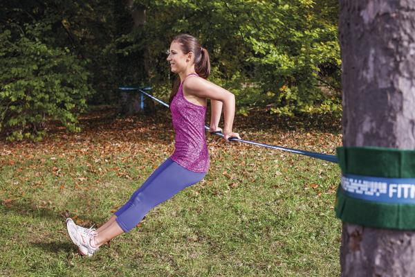 Frau trainiert mit Fitnessband an Baum im Park, Ganzkörper-Workout im Freien.