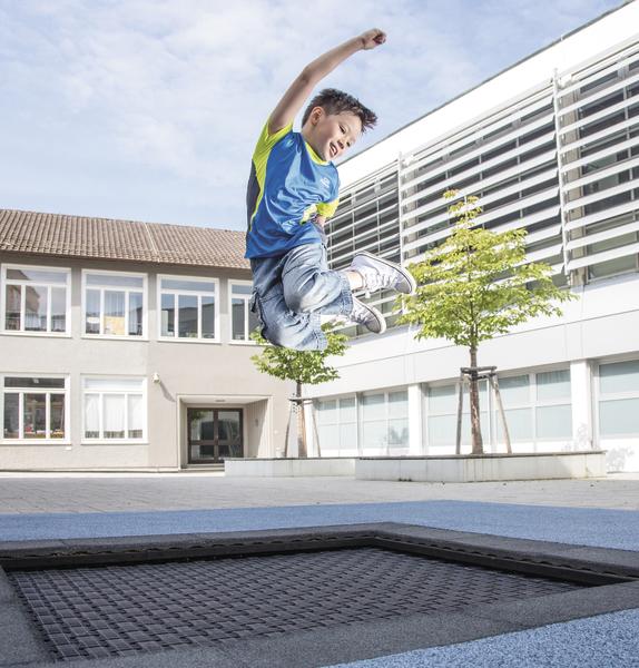Junge springt fröhlich auf einem Trampolin im Schulhof bei sonnigem Wetter.
