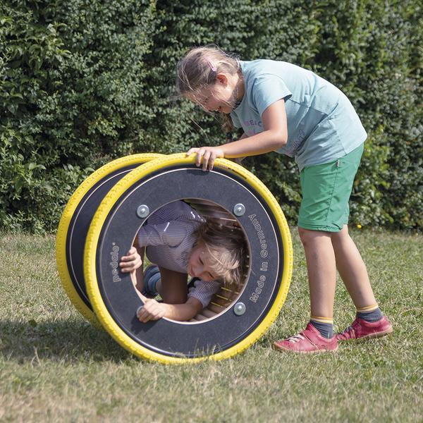 Kind spielt draußen mit großem gelb-schwarzem Spielrad auf Grasfläche vor Hecke.
