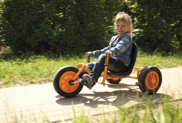 Kind fährt fröhlich auf einem orangefarbenen Dreirad auf einem Weg im Grünen.