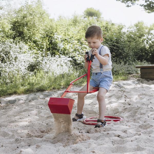 Kleiner Junge spielt mit rotem Sandbagger im Sandkasten an sonnigem Tag im Freien.
