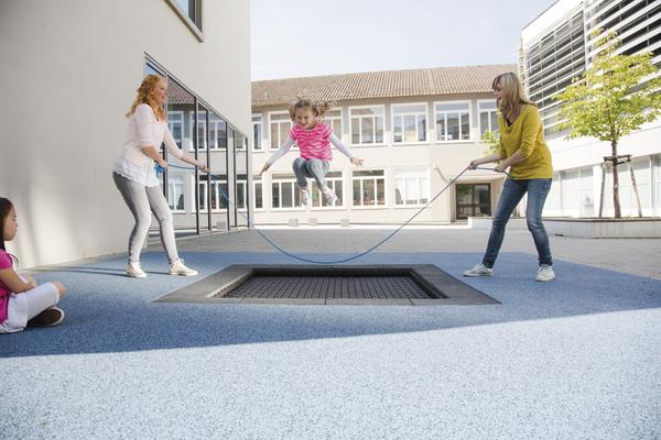 Kind springt auf eingebautem Trampolin, zwei Frauen schwingen Springseil im Schulhof.