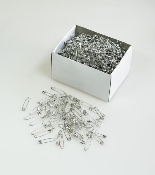 Weiße Schachtel mit vielen silbernen Sicherheitsnadeln, einige daneben liegend, auf weißem Hintergrund.