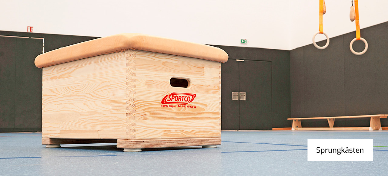 Ein Sprungkasten aus hellem Holz von der Marke Sportco steht in einer Sporthalle. Im Hintergrund sind Turnringe und eine Turnbank zu erkennen. Unten rechts ist ein Textfeld mit der Aufschrift "Sprungkästen".