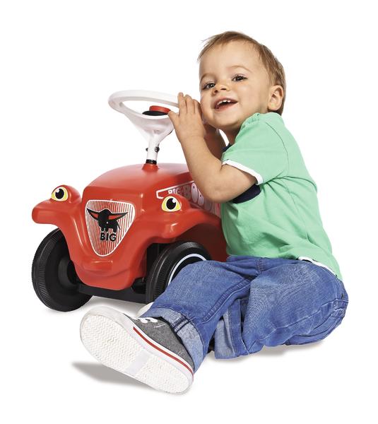 Kleinkind sitzt lachend neben rotem Rutschauto mit Lenkrad auf weißem Hintergrund.