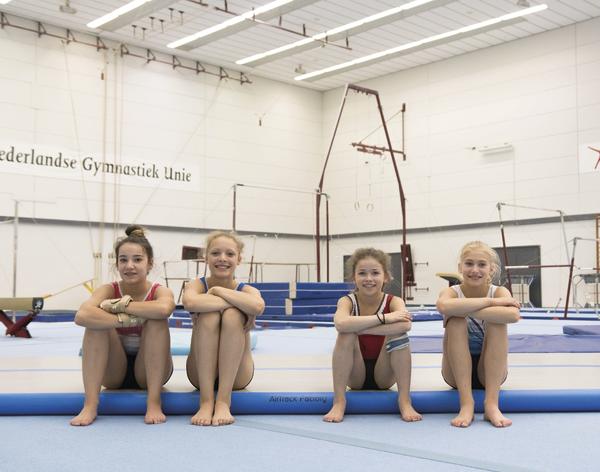 Vier junge Turnerinnen sitzen auf einer Gymnastikmatte in einer Turnhalle.