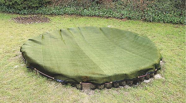 Abgedecktes rundes Erdloch mit grünem Schutznetz auf einer Rasenfläche.