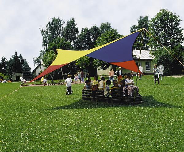 Kinder sitzen unter einem bunten Sonnensegel auf einer grünen Wiese im Freien.
