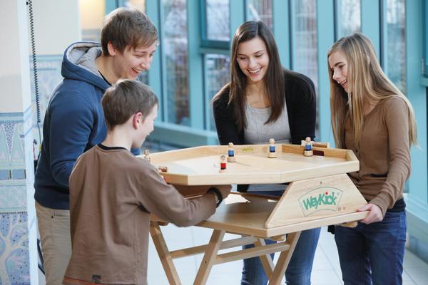 Vier Jugendliche spielen gemeinsam an einem Holz-WeyKick-Tischspiel in hellem Raum.