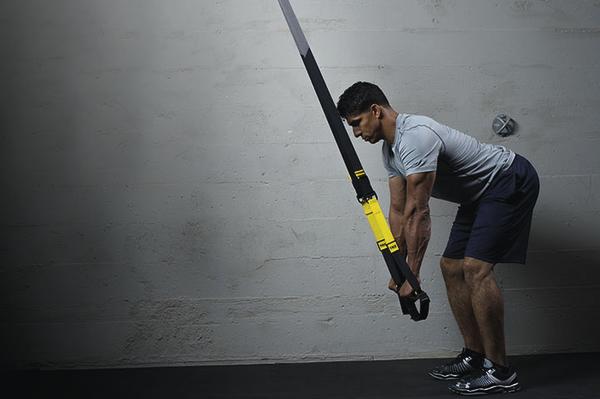 Mann trainiert mit TRX-Bändern in Fitnessstudio vor grauer Wand, Ganzkörperübung.