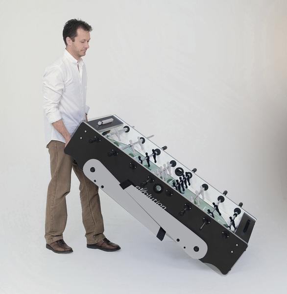 Mann hebt und trägt einen zusammengeklappten Tischkicker in einem weißen Raum.