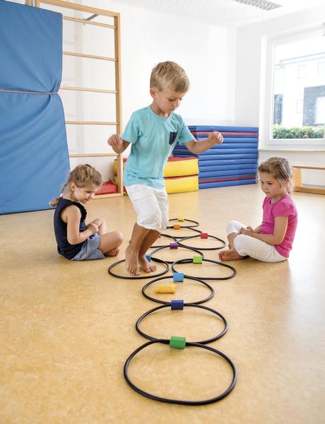 Kind spielt in Turnhalle mit Hula-Hoop-Reifen und bunten Blöcken, zwei Kinder sitzen daneben.