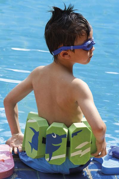 Junge mit Schwimmgürtel und Schwimmbrille sitzt am Poolrand im blauen Wasser.