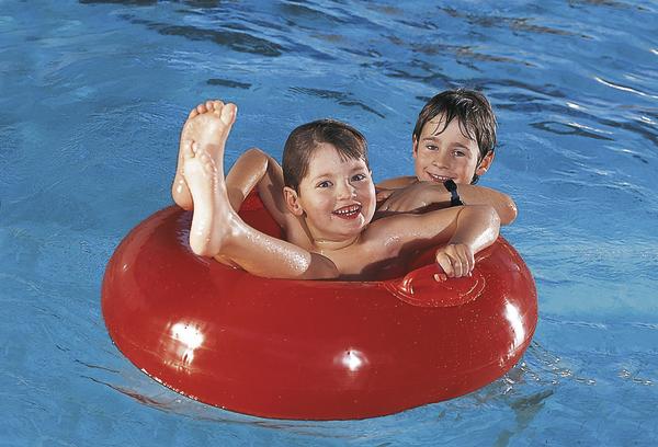 Zwei lachende Kinder spielen im Schwimmbad auf einem roten Schwimmreifen.