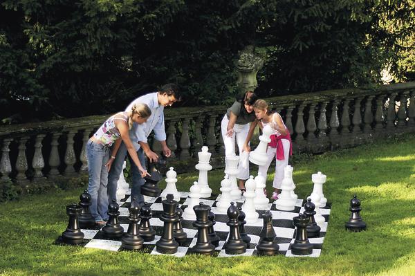 Vier Personen spielen im Garten Schach mit übergroßen Figuren auf einem großen Schachbrett.