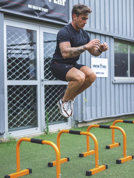 Mann trainiert draußen mit Sprüngen über orangefarbene Hürden auf Kunstrasen.
