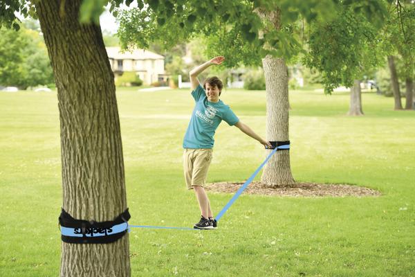 Junge balanciert auf einer blauen Slackline zwischen zwei Bäumen im Park bei Sonnenschein.